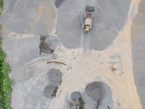 Wheel Loader Surrounded by a Large Aggregate Stockpiling Area in Indonesia. Heavy Construction Machinery Stands Among Rock, Sand, and Processed Materials in Industrial Quarry Operations from Above.