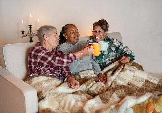 Happy multiracial senior women cheering with hot tea sitting on couch during winter vacation - Female friendship, elderly people and lifestyle concept