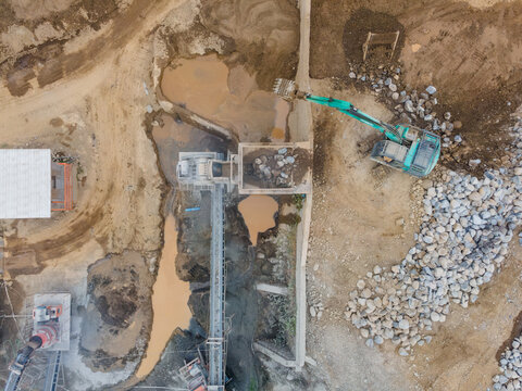 Mining Work in a Rock and Sand Quarry with an Excavator near Rock Crusher Machine Working on a Pile of Large Rocks. Heavy Equipment Operates Across the Active Mining Site from Above.