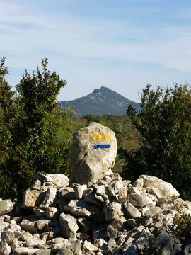 A cairn stone with yellow and blue trail paint markings in the foreground, framing the distinct peak of Pic Saint-Loup mountain under a clear sky.