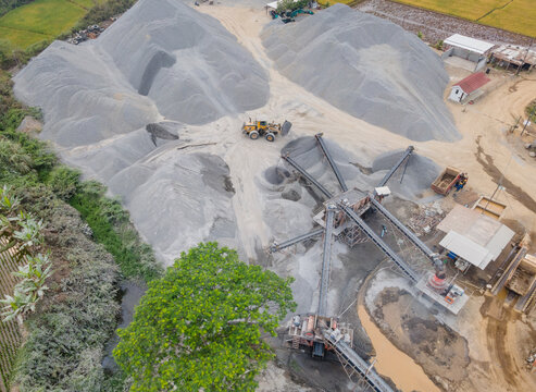 Wheel Loader and Conveyor Belt Transporting Split Rocks in the Middle of an Aggregate Stockpiling Area. Heavy Machinery Operates Among Rock, Sand, and Processed Materials Viewed from Above.