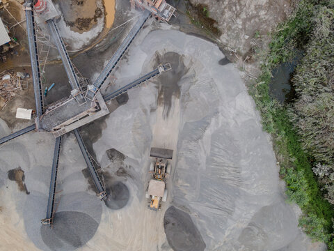 Wheel Loader and Conveyor Belt Transporting Split Rocks in the Middle of an Aggregate Stockpiling Area. Heavy Machinery Operates Among Rock, Sand, and Processed Materials Viewed from Above.