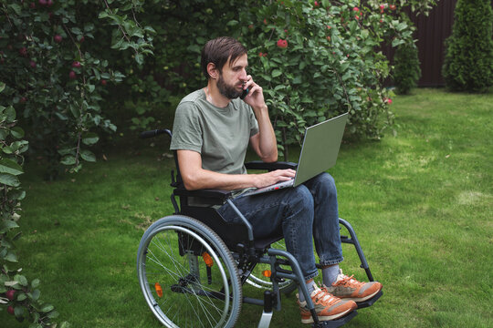 Bearded man in wheelchair working on laptop, talking on phone on green lawn in garden with apple trees, concept of remote work for people with disabilities, inclusion, freelance, equal opportunity