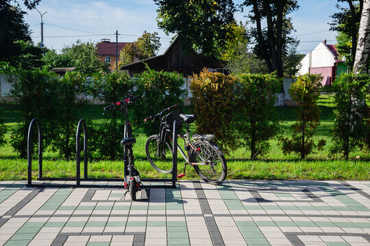Bicycle and electric scooter parked at metal rack on tiled pavement of outdoor public area near green hedge
