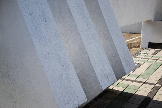 Massive white concrete structural pillars casting rhythmic geometric shadows on tiled outdoor pavement of public building exterior