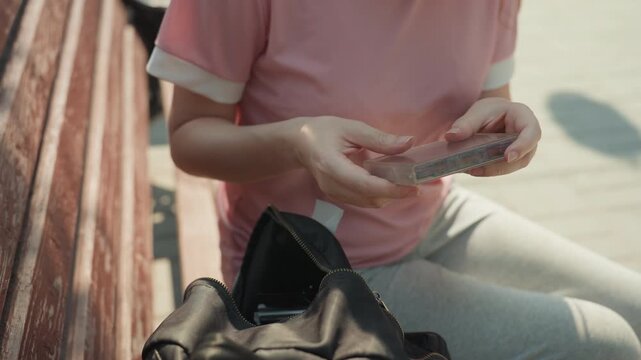 Young woman in pink shirt examines and stores cassette tapes while sitting on wooden bench. Woman handles transparent cases with visible magnetic tape reels placing them into open black bag