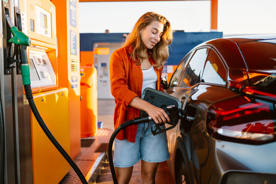 Smiling woman stands by her car at a gas station on a sunny day. The driver is refueling the vehicle and enjoying the moment. A concept of transportation, fuel, and energy efficiency.