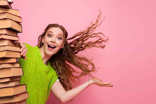 Young woman with green sweater laughs and hugs stack of book against pink background conveying study fun education energy and playful confidence