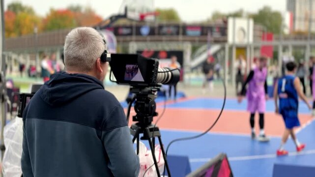 Moscow, Russia - September 27, 2026: Video operator records players moving across a colorful outdoor basketball court while spectators watch from surrounding areas