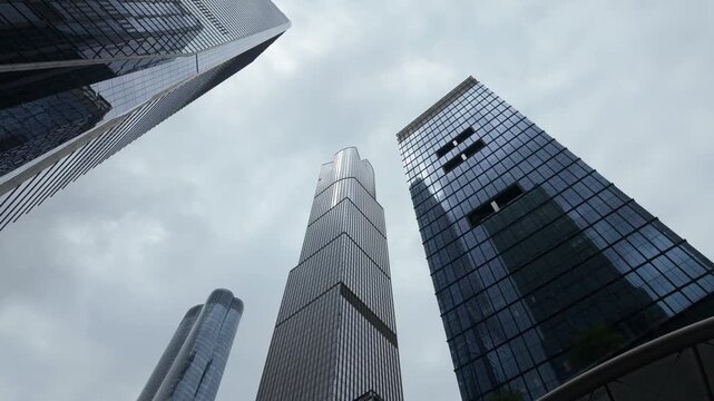 Low-angle view of modern skyscrapers with glass facades in a financial district revealing green trees at the base, representing business, economy, and corporate architecture