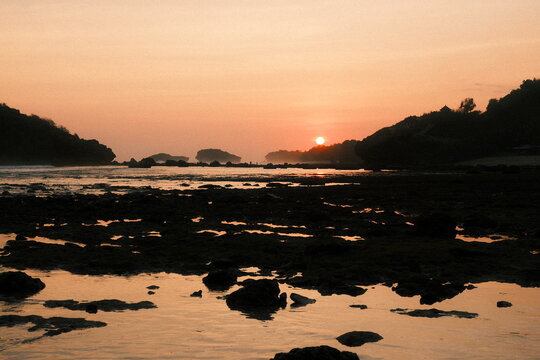 Sun setting behind silhouetted hills over a rocky tidal beach