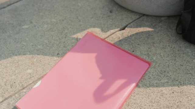 Hand places pink folder on concrete then adds transparent cassette tape and pen. scene features soft shadows from objects in outdoor setting with natural light