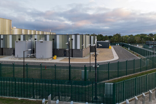 Aerial view of a data center with industrial buildings, large storage tanks, and a secure perimeter fence with surveillance cameras under a cloudy sky in the United Kingdom.