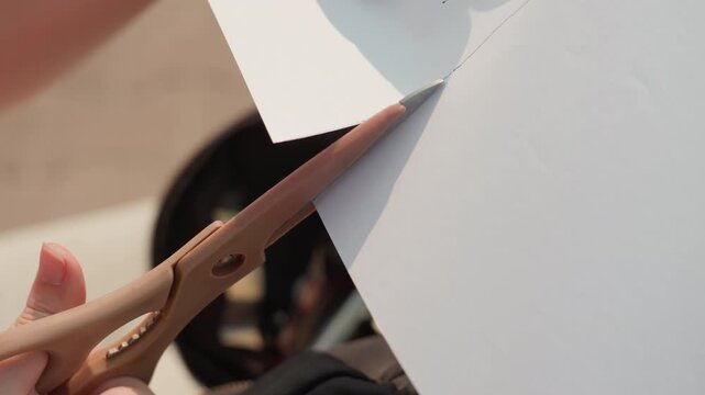 Hand carefully cuts white sheet of paper with brown scissors focusing on precise movement and interaction between tool and material. scene is set outdoors under natural light emphasizing