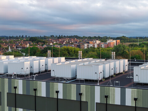 Aerial view of a data center rooftop featuring rows of white cooling units and infrastructure, with residential buildings and trees in the background in the United Kingdom.