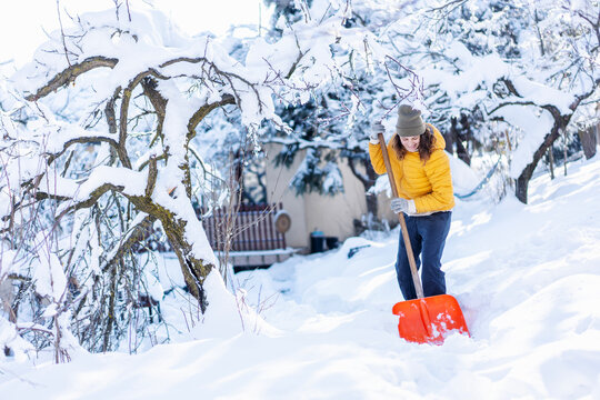  Young caucasian woman in yellow jacket shoveling snow from the path in a country house yard.