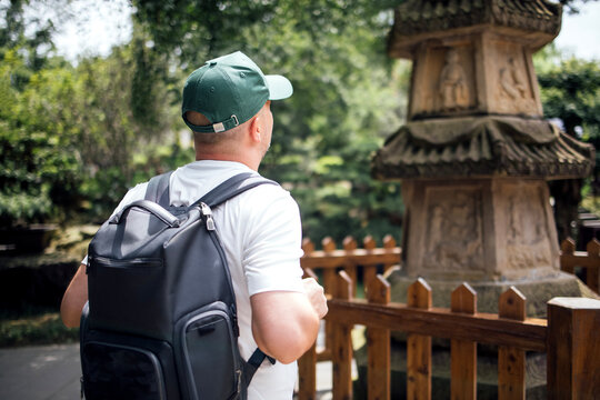 Male tourist wearing a green cap and white shirt stands near a stone structure in a garden, with wooden fence and lush greenery in the background