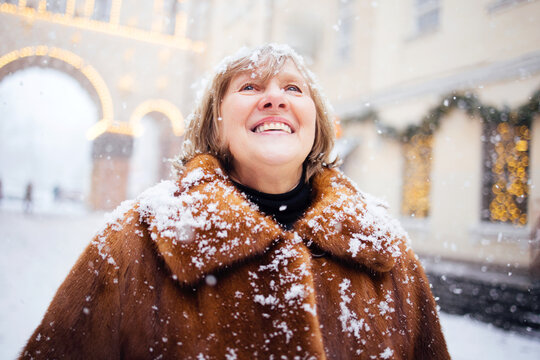 Middle-aged woman with short blonde hair wearing a brown fur coat stands outdoors in falling snow, smiling while looking up at the winter sky in a festive city setting