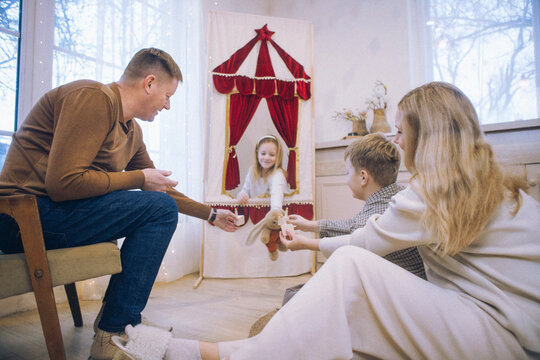 Family enjoying a playful moment with a girl in a puppet theater, seated on a chair while two children engage in imaginative play in a cozy indoor setting