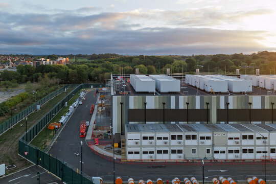 Aerial view of a large data center facility with modular office units and construction equipment, surrounded by a security fence and lush green trees in the United Kingdom.