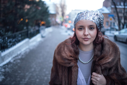 Young woman with red hair wearing a sequined headband and fur coat poses confidently on a snowy street with blurred city lights in the background