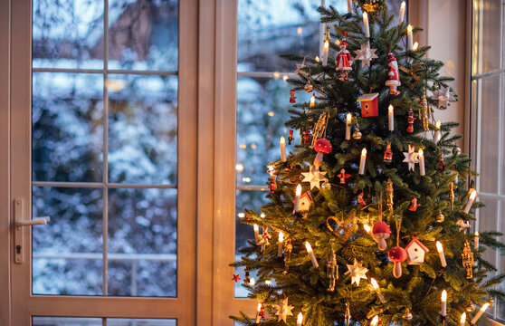 Christmas tree adorned with colorful ornaments and warm lights, standing near a window with a snowy landscape outside, creating a cozy holiday atmosphere