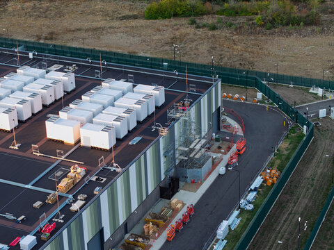 Aerial view of a data center facility featuring rows of white modular units, a large building with green and grey panels, scaffolding, and a fenced perimeter in the United Kingdom.