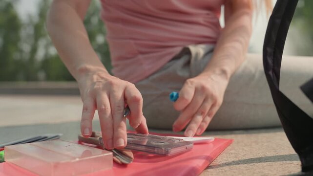 Young woman in pink shirt organizes stationery items like pens scissors and cassette tapes on red folders outdoors. Woman arranges objects including transparent plastic containers