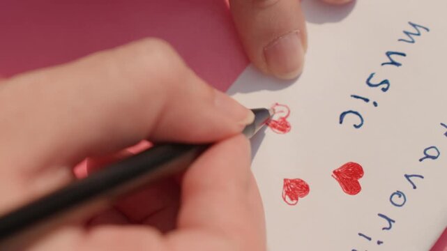 Hand holding pen draws red hearts on paper with blue handwritten text including music and favors. scene is bright indoor lighting focusing on close up of hands and drawing activity