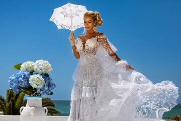 Elegant Blonde Bride in Couture Lace Wedding Gown with Flying Veil and Parasol, Posing by Azure Sea with Blue Hydrangeas at a Luxury Mediterranean Coastal Resort © marcink3333