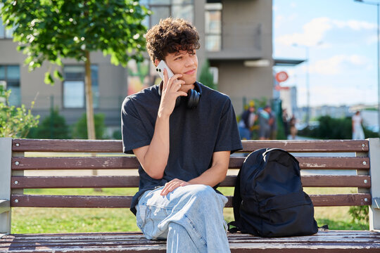 Young male student talking on smartphone sitting on bench outdoors in park