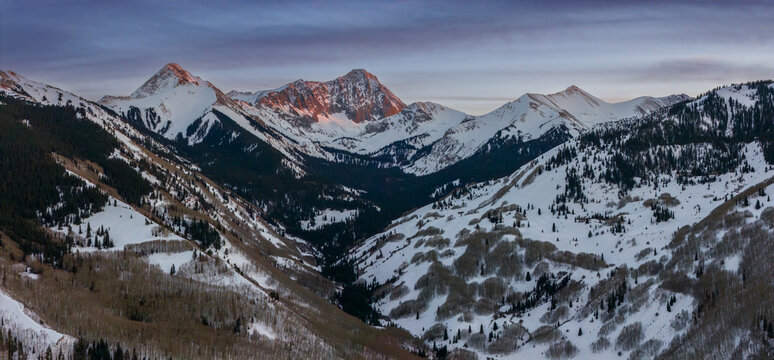 Aerial view of Capitol Peak in the Rocky Mountains during a winter sunset, featuring snow-covered slopes and evergreen forests in Aspen, Colorado, United States.