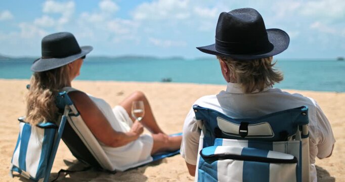 Senior couple sitting on beach chairs enjoying a tropical vacation by the ocean, man pointing towards the horizon, woman holds glass of wine, embracing leisure and traveling to Samui Island, Thailand