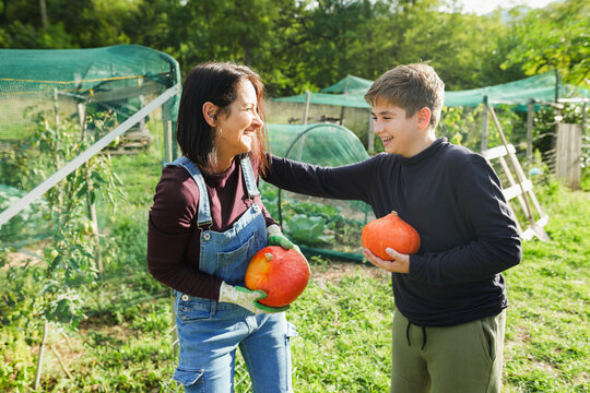 Mother and son holding organic pumpkins with hands inside vegetable home garden - Family people having fun during fall season - Autumn lifestyle concept - UGC wide lens content