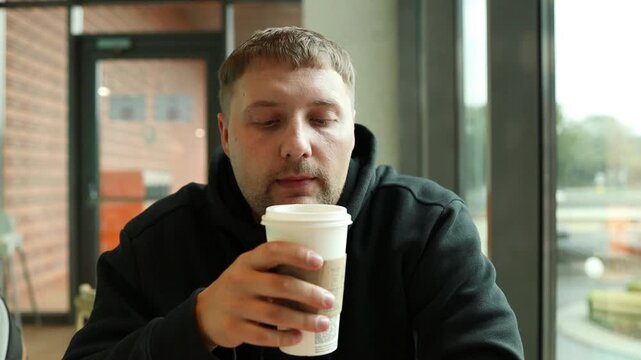 Detailed horizontal shot of a young caucasian man in a black hoodie sipping a warm beverage from a disposable takeaway cup inside a modern restaurant or cafeteria. 