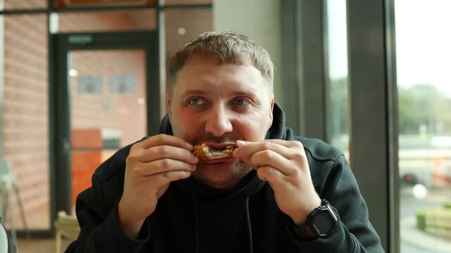 Close-up portrait of a young caucasian man enjoying a delicious piece of crunchy deep-fried chicken wing or strip inside a cozy modern cafe.