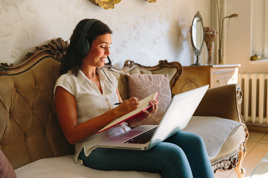 Woman taking notes while using laptop and headphones at home