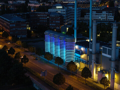 Aerial view of the BTB Heat and Power Plant Adlershof with glowing blue and purple light installations on the storage tanks at night in Berlin, Berlin, Germany.