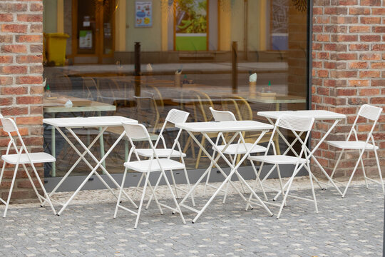 Outdoor cafe tables and white folding chairs arranged on a cobblestone sidewalk beside a brick storefront with reflective windows, urban street seating