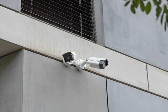 Two outdoor security cameras mounted on a modern concrete building facade beneath a dark louvered window with foliage at the edge, architectural surveillance equipment