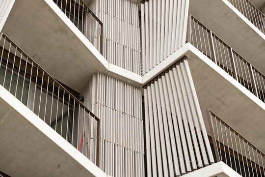 Geometric apartment building facade with concrete balconies, metal railings, and vertical white architectural screens viewed from below in an urban residential structure
