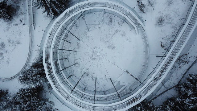 Aerial view of the Col de la Faucille summer sledge track covered in snow, featuring a circular metal structure surrounded by snow-covered pine trees in Gex, Auvergne-Rhone-Alpes, France.