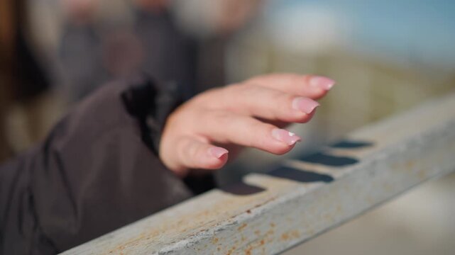 Woman hand with manicured nails rests on weathered metal railing showing subtle movement and texture detail. background is softly blurred suggesting outdoor setting like bridge or deck