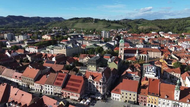 Aerial view of historic Maribor, Slovenia, showing the old town, red rooftops, cathedral tower, surrounding green hills and vineyards under a clear blue sky.