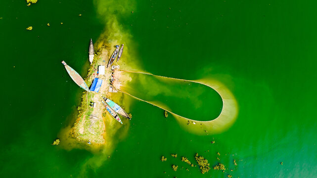 Aerial view of a small island in Rangamati with wooden fishing boats and a large net being cast into the vibrant green lake water, Rangamati, Chittagong Division, Bangladesh.