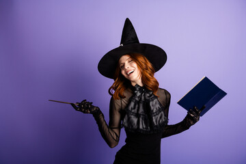Naklejka na ściany i meble Smiling redhead witch in black hat and dress holding a magic book and wand against a purple background, playful Halloween costume portrait