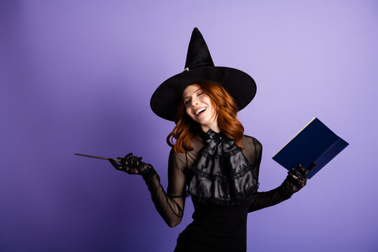 Smiling redhead witch in black hat and dress holding a magic book and wand against a purple background, playful Halloween costume portrait