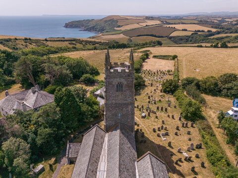 Aerial view of the historic stone church and its graveyard surrounded by lush green fields and trees with the scenic coastline in the background in Lansallos, England, United Kingdom.