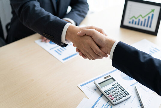 A handshake between two business professionals at a table, with a calculator and financial charts visible, signifying a successful negotiation and agreement on financial matters.