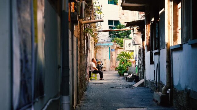 Man sitting on a stool in a narrow sunlit alley with weathered walls and warm light